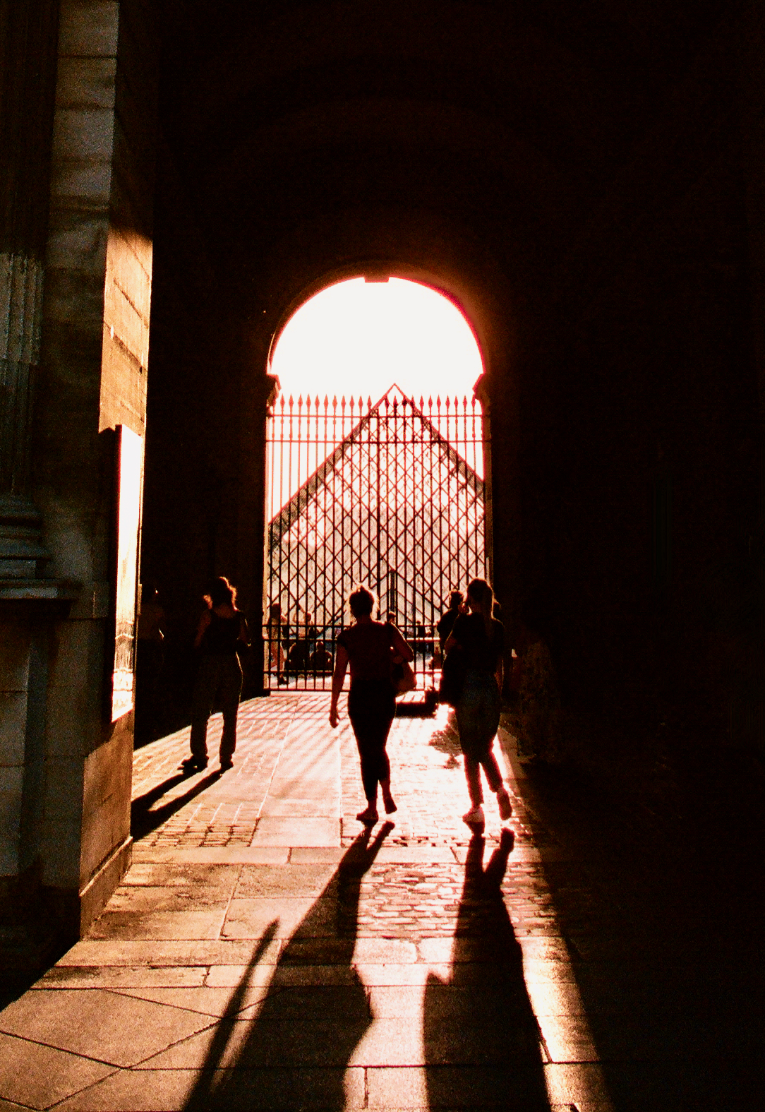 People silhouetted in an archway with the louvre pyramid beyond the gate.